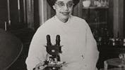 Image of woman with dark hair wearing glasses and a laboratory coat sitting with a microscope in front of her.  There is a display case behind her.